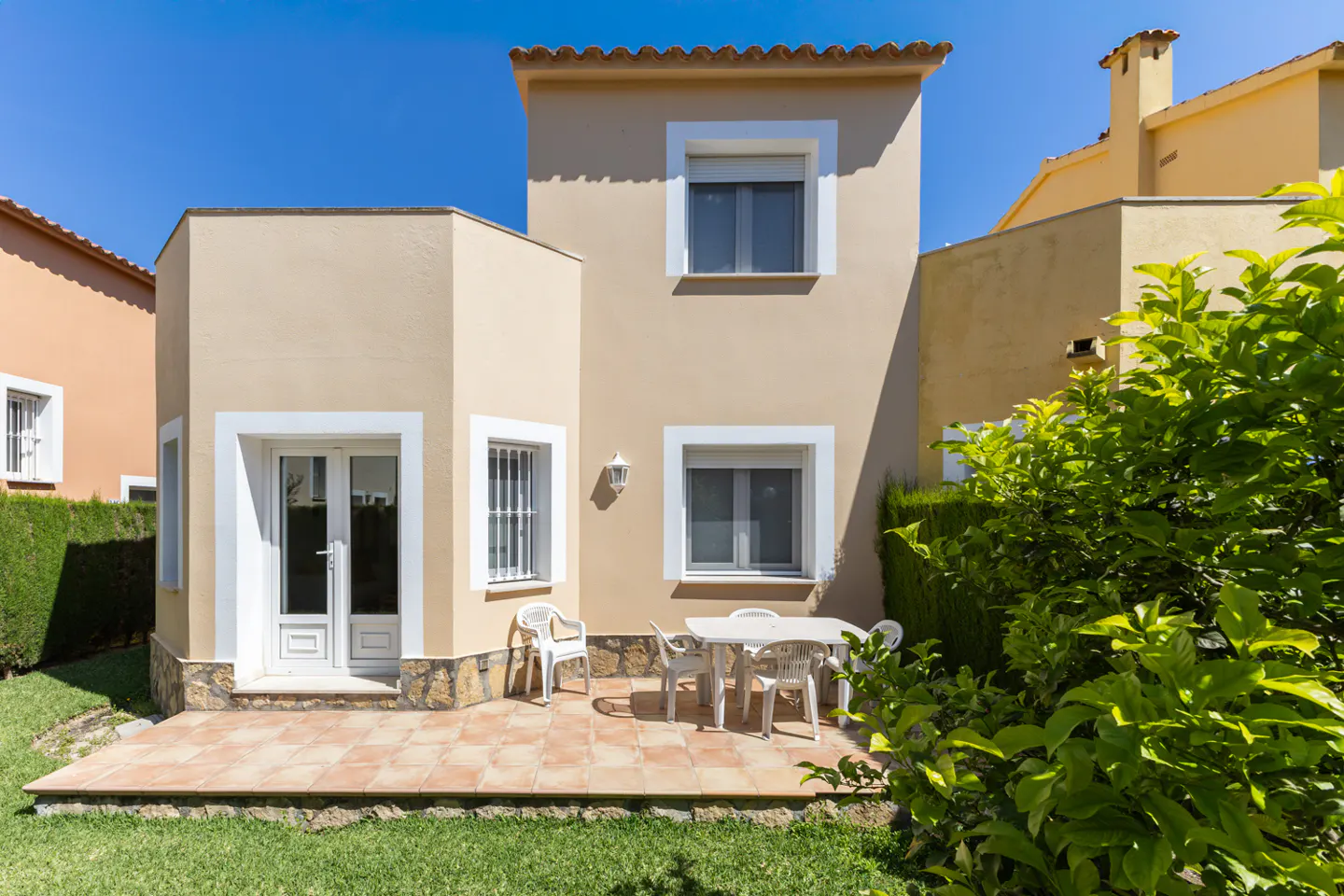 Exterior view of a two-story beige house with a patio, table, chairs, and green lawn under a clear blue sky.