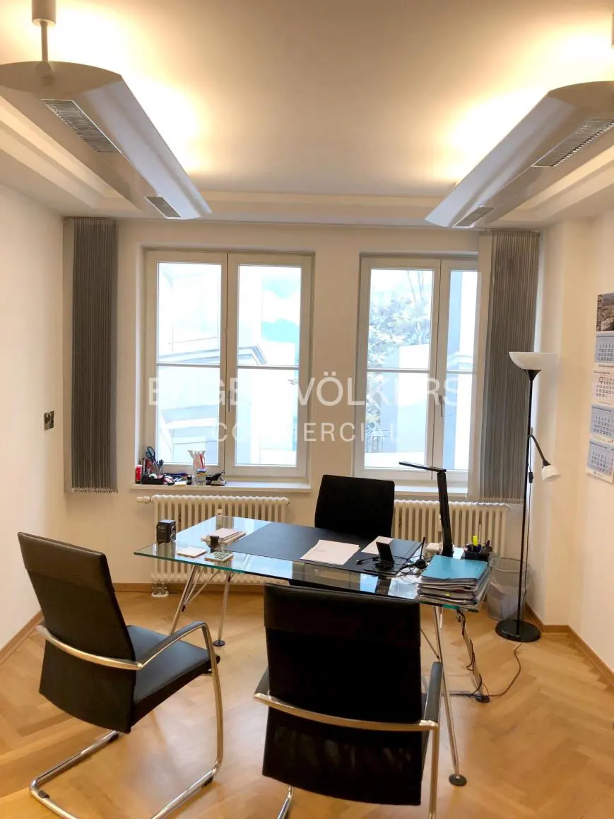 Office space with a glass desk, black chairs, and herringbone wood floors. Windows provide natural light.