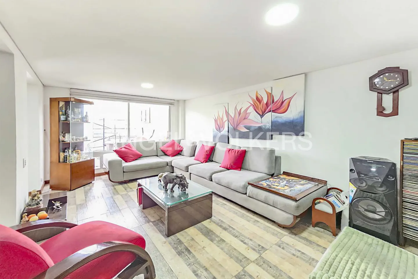 Bright living room with gray sectional sofa, red pillows, glass coffee table, wood floors, and large floral art.