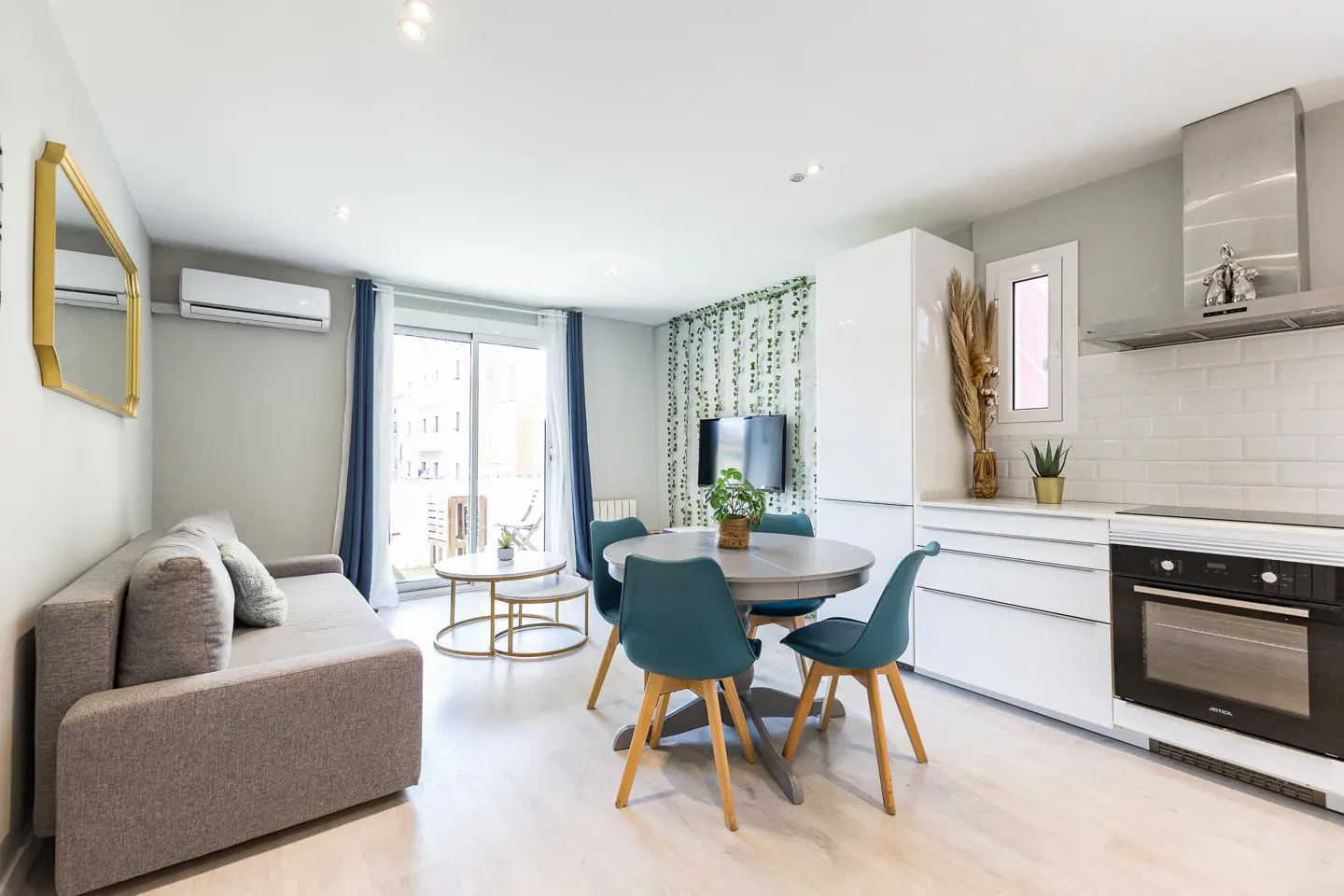 Bright, modern apartment interior with a gray sofa, round table with teal chairs, and a white kitchen area.