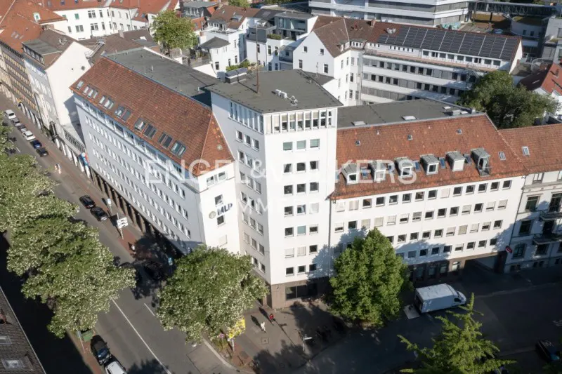 Aerial view of a white office building with a red-tiled roof, surrounded by trees and other buildings in a city.