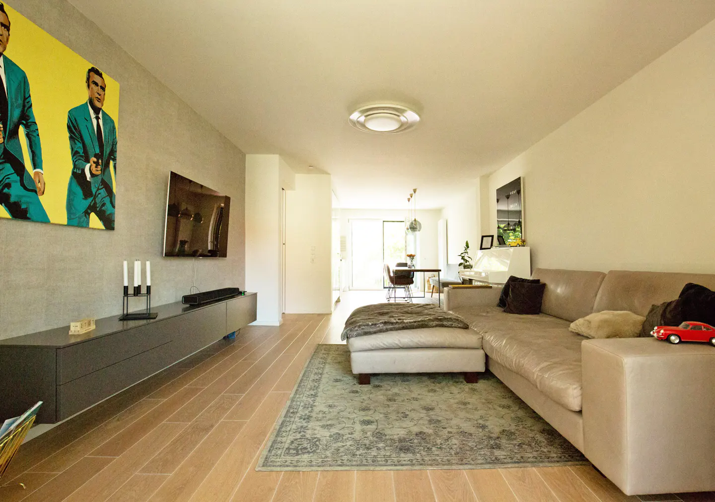 Bright living room with a beige sectional sofa, a gray media console, and a large pop art painting. Wood floors and a patterned rug add warmth.