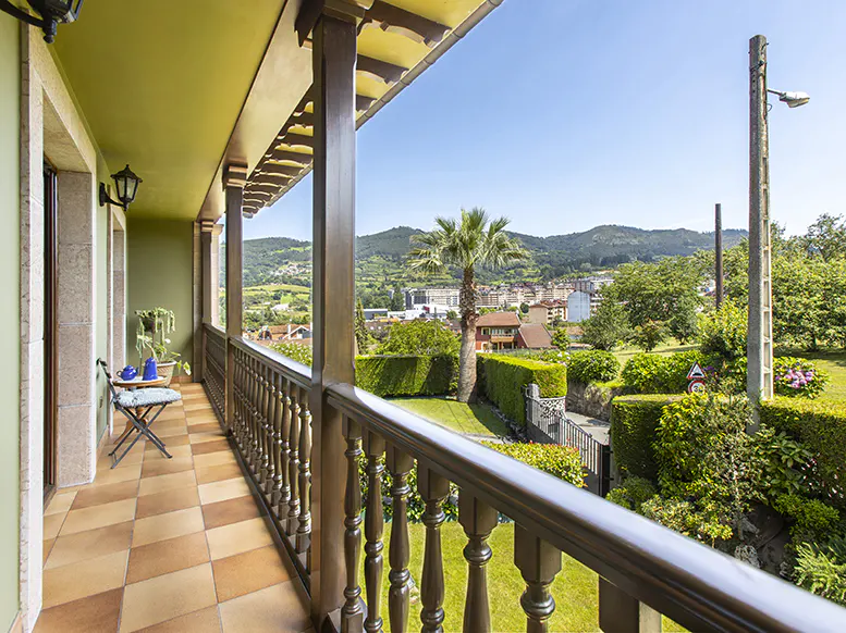 Balcony view with brown railings, checkered tile floor, and a chair. A green landscape with houses and mountains is visible in the background.