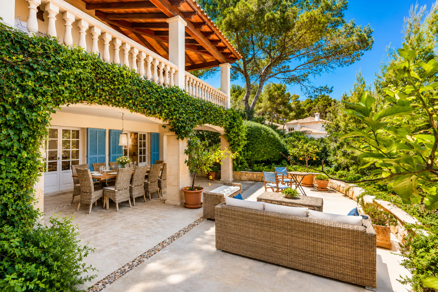 A sunny Mediterranean patio with a vine-covered villa, outdoor dining set, and wicker lounge furniture in a lush garden.