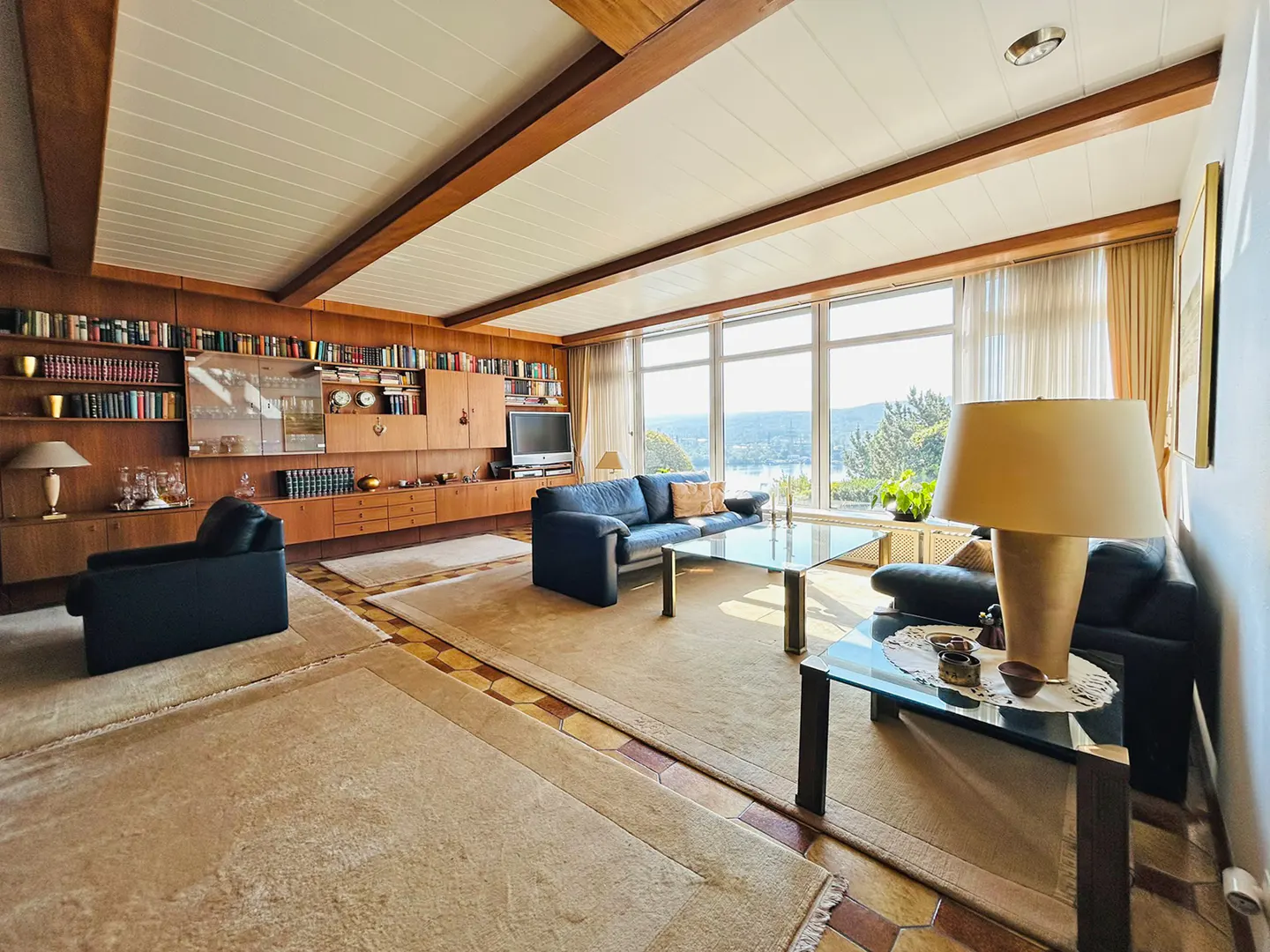 A living room with a large window, blue sofas, glass tables, and a built-in bookshelf. The room has a beige carpet and a white ceiling with wooden beams.