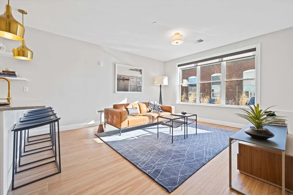 Bright living room with a brown leather sofa, blue rug, and wood floors. Large window with a view of a brick building.