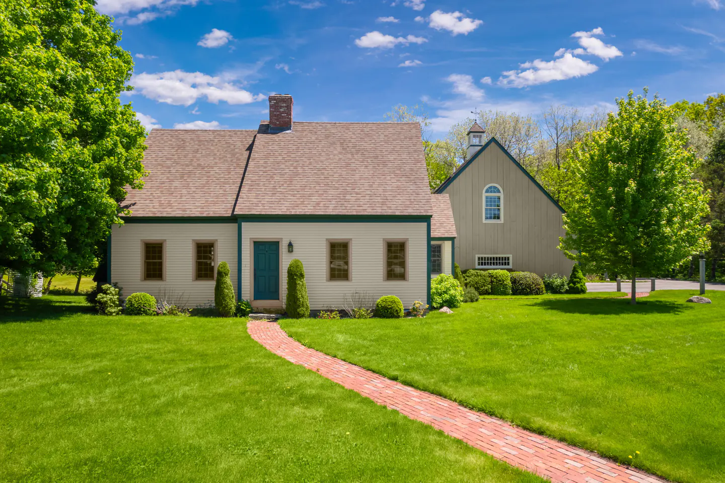 A charming light-colored home with a brick walkway, green lawn, and a matching barn under a bright blue sky.