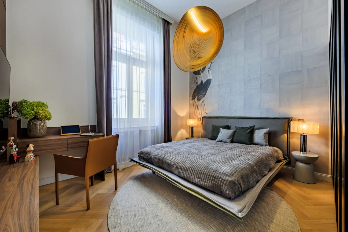 Bedroom with a gray bed, round rug, desk, and a large, round, gold pendant light. A crane mural is on the wall behind the bed.