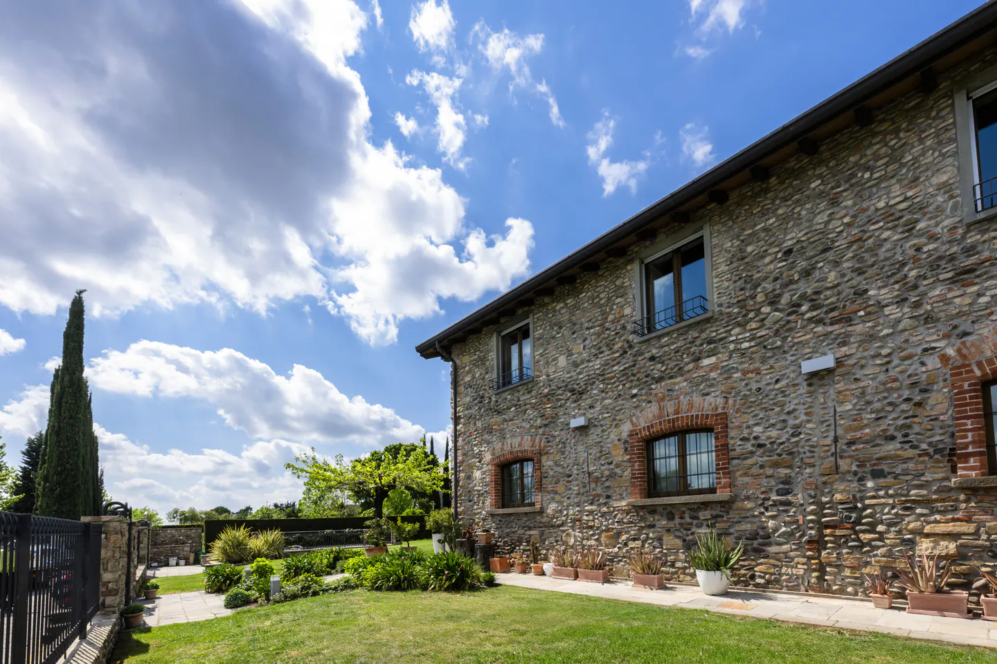 Exterior view of a stone house with a green lawn, potted plants, and a blue sky with white clouds.