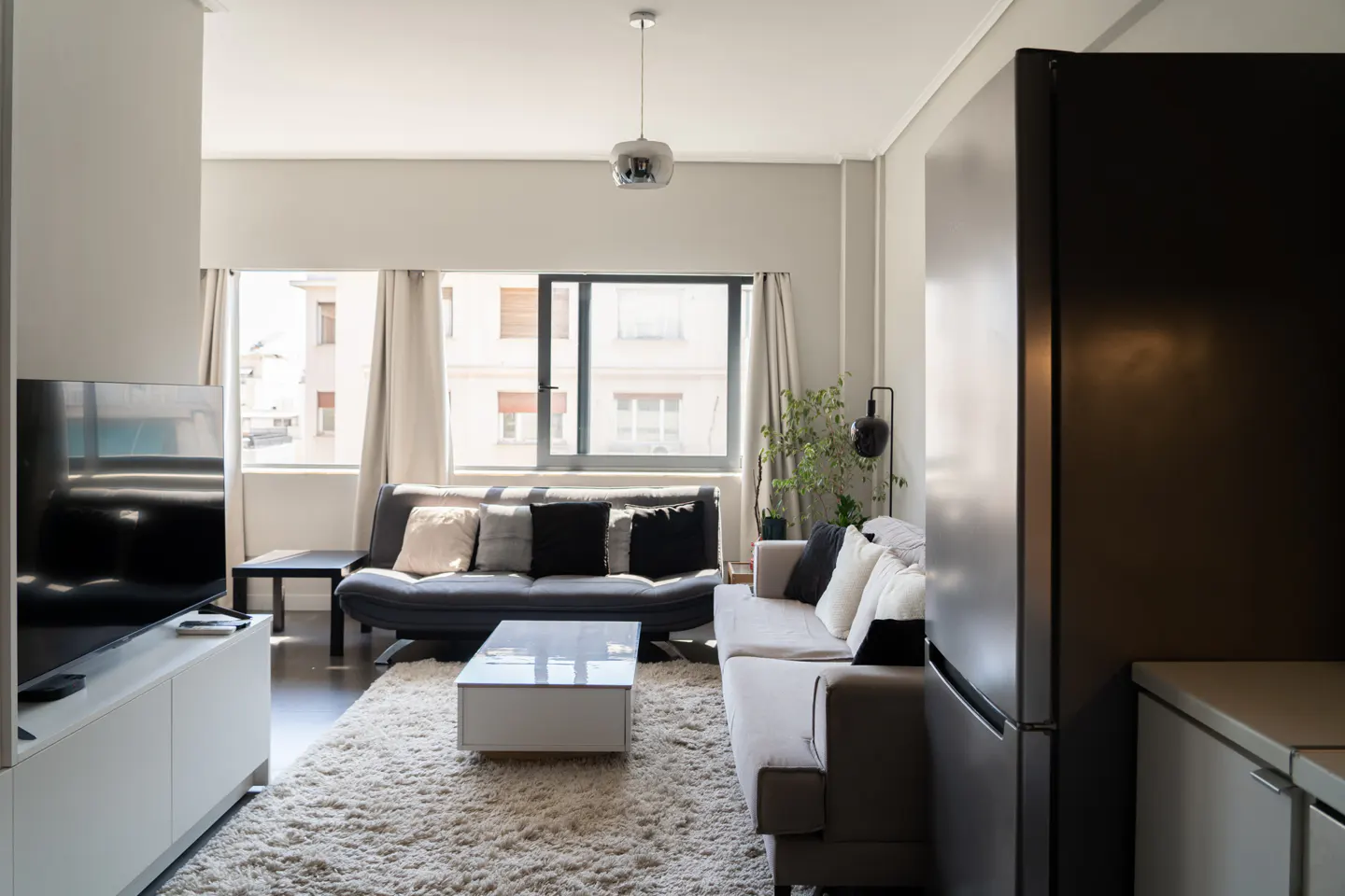 Modern living room with a grey sofa, white coffee table, shag rug, and a large stainless steel refrigerator.