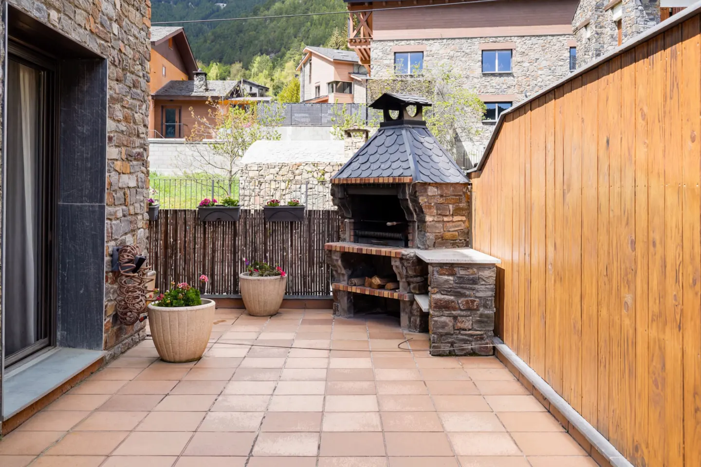 Outdoor patio with stone grill, wood fence, and tile floor. Flower pots add color. Houses and mountains are visible in the background.