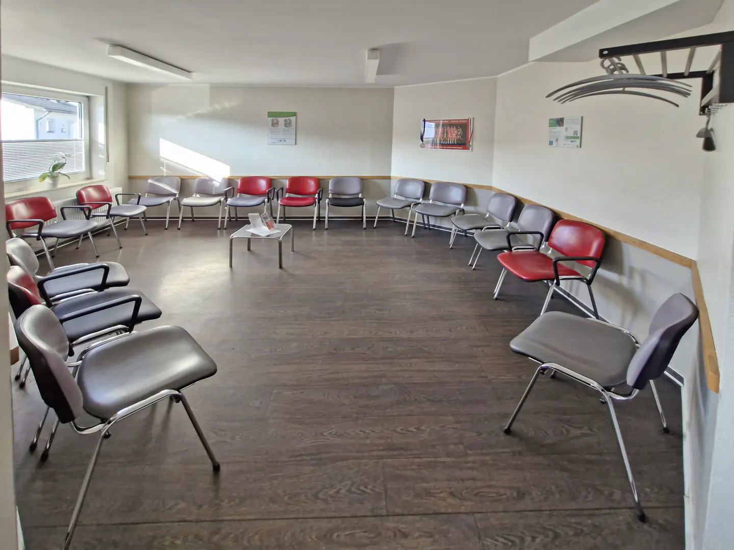 A waiting room with red and grey chairs arranged in a circle on a dark wood floor, with a small table in the center.