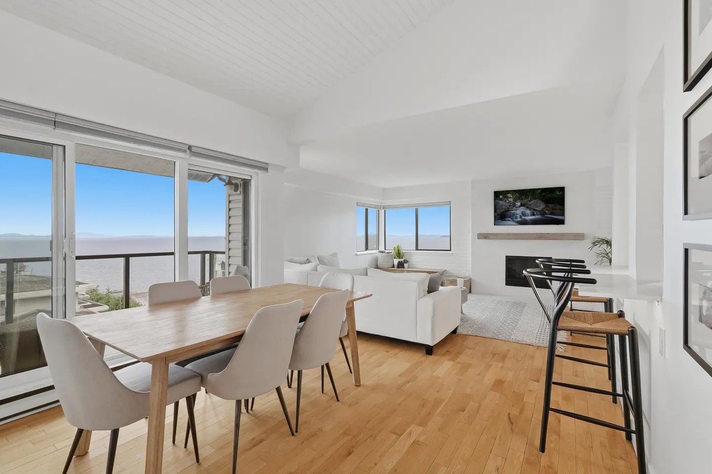 Bright, open-concept living space with wood floors, dining table, white sofa, fireplace, and ocean view.