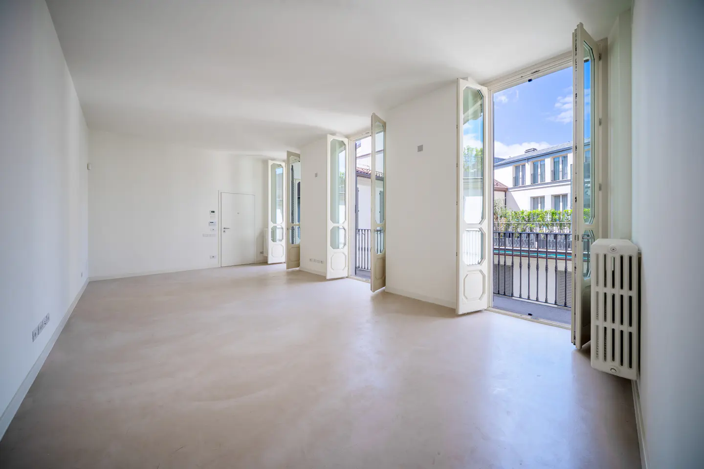 Bright, empty room with white walls and light floor. Three sets of open, white French doors lead to balconies with black railings.
