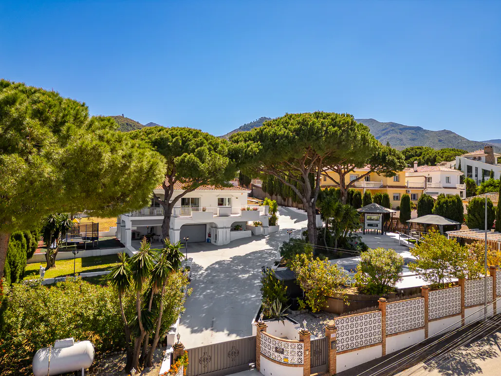 Aerial view of a white villa with a gray driveway, surrounded by green trees and a decorative fence under a clear blue sky.