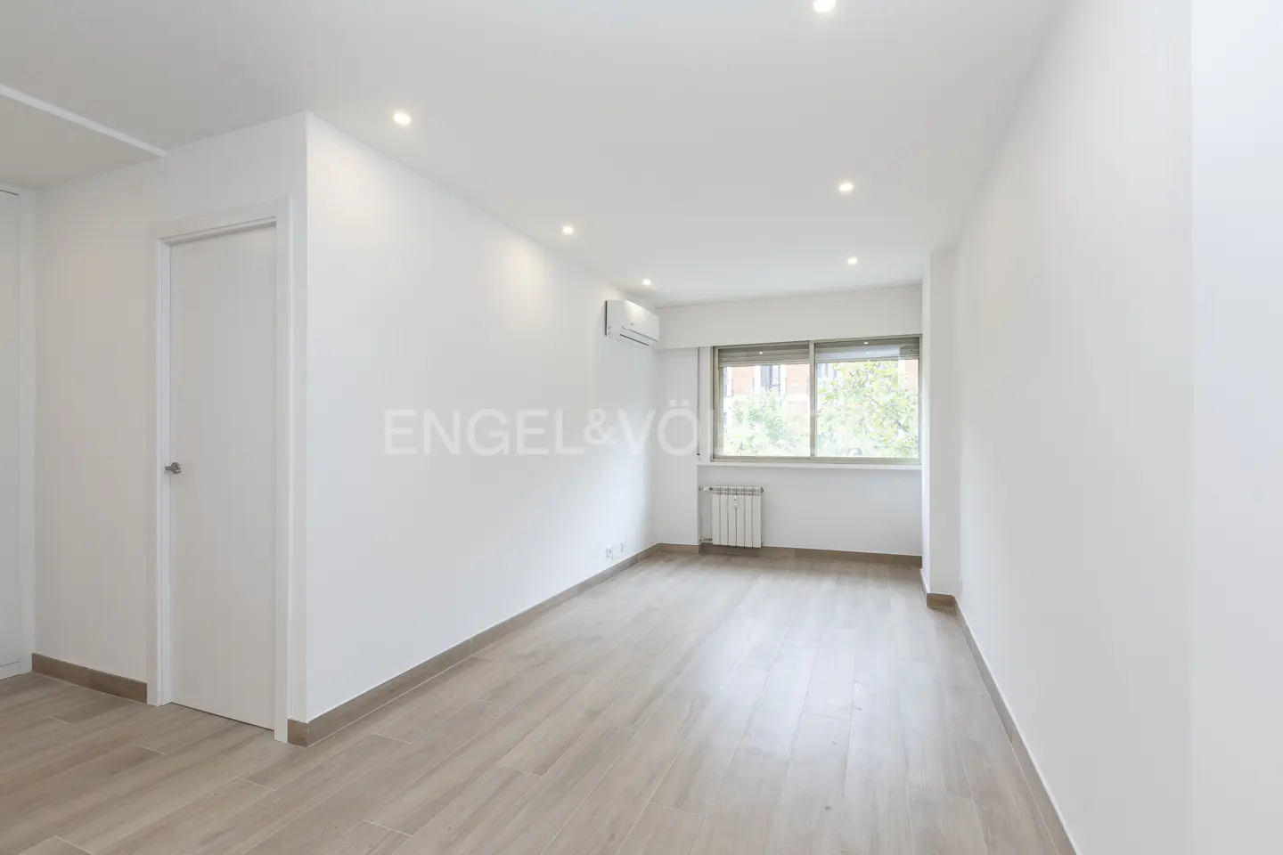 Bright, empty room with light wood floors, white walls, and a window. A white door is on the left, and the Engel & Völkers logo is on the wall.