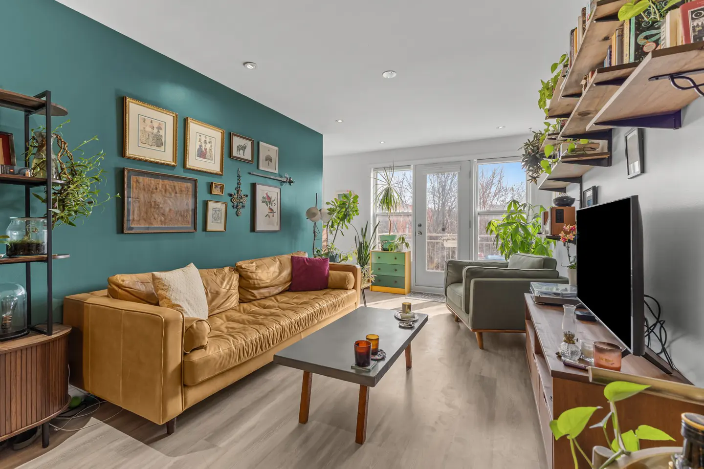 Bright living room with a tan leather sofa, teal accent wall with framed art, and a gray coffee table. Plants add a touch of nature.