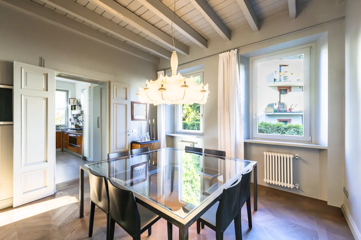 Dining room with a glass table, black chairs, and a modern chandelier. White curtains frame the windows, and a glimpse of the kitchen is visible through an open doorway.