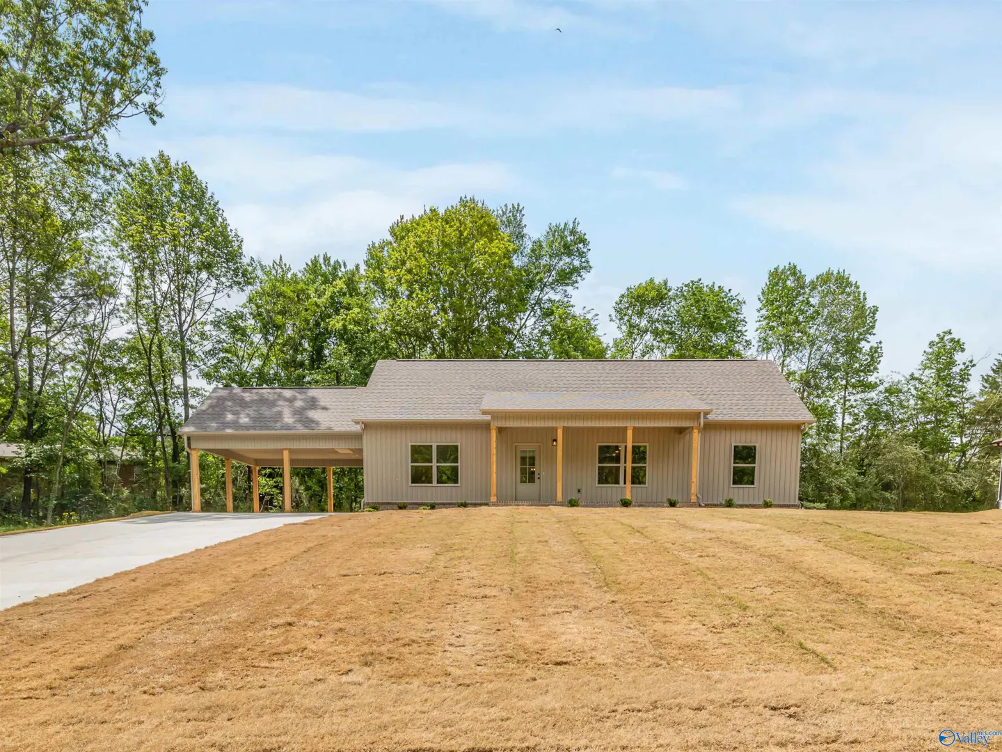 A single-story beige house with a carport and porch sits on a brown lawn under a blue sky.