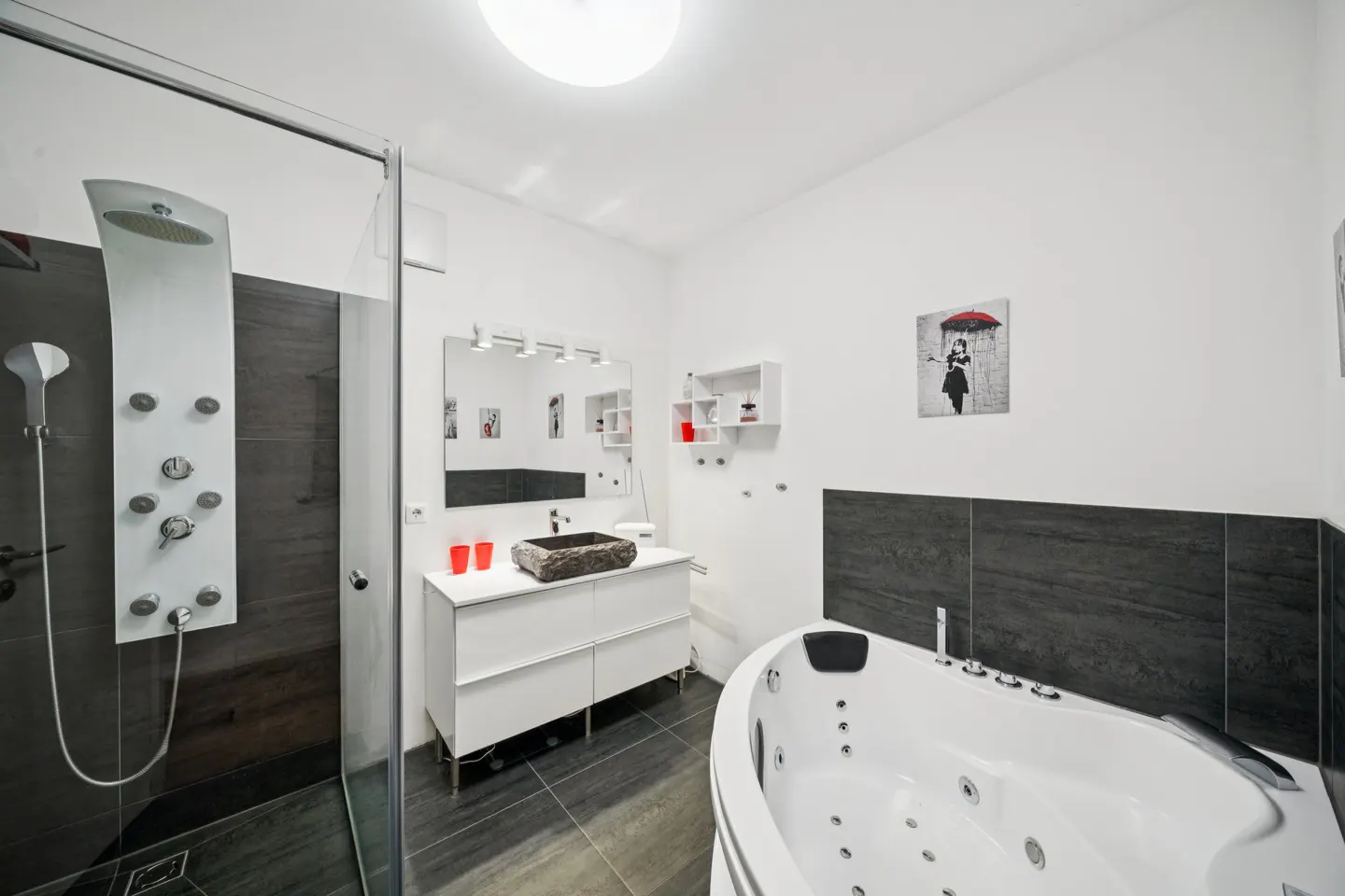 Modern bathroom with a glass shower, white vanity with a stone sink, and a white jacuzzi tub against a gray tiled wall.