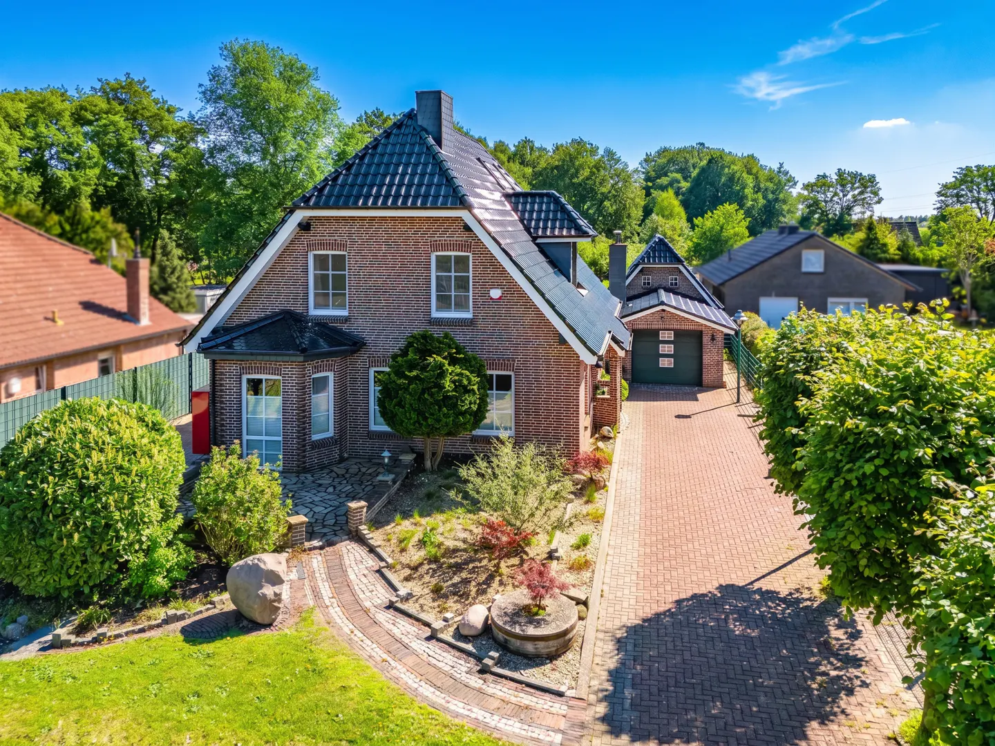 Aerial view of a brick house with a dark roof, green lawn, and brick driveway on a sunny day.