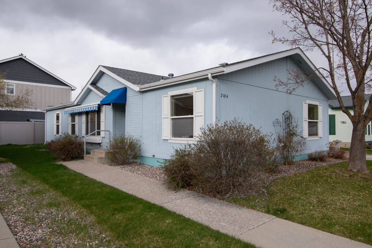 A light blue single-story house with white trim and a blue awning over the front door. A concrete walkway leads to the entrance.