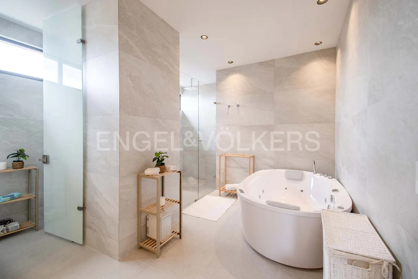 Bright bathroom with marble walls, a glass shower, and a white jacuzzi tub. Wooden shelves hold plants and towels.