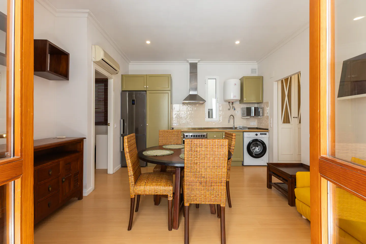A bright kitchen with a round table and four wicker chairs. Olive green cabinets, stainless steel appliances, and a washing machine are visible.