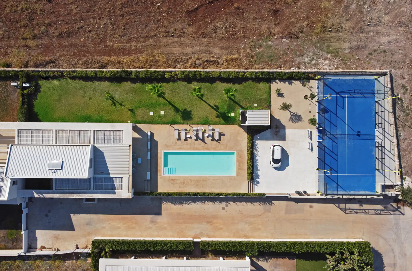 Aerial view of a modern villa with a pool, tennis court, and manicured lawn. A white car is parked nearby.