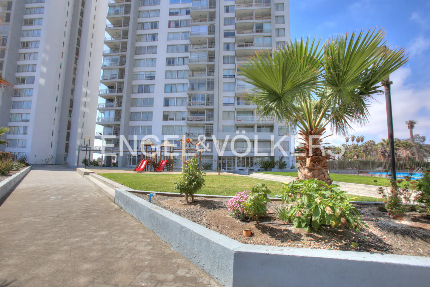 Exterior view of a tall, white apartment building with balconies, a playground, and palm trees on a sunny day.