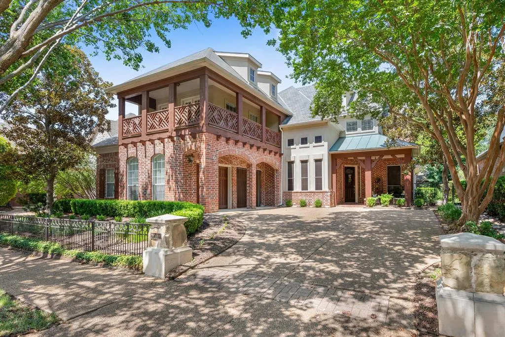 Two-story brick home with a balcony and arched garage doors. A stone driveway leads to the front door, framed by green trees.