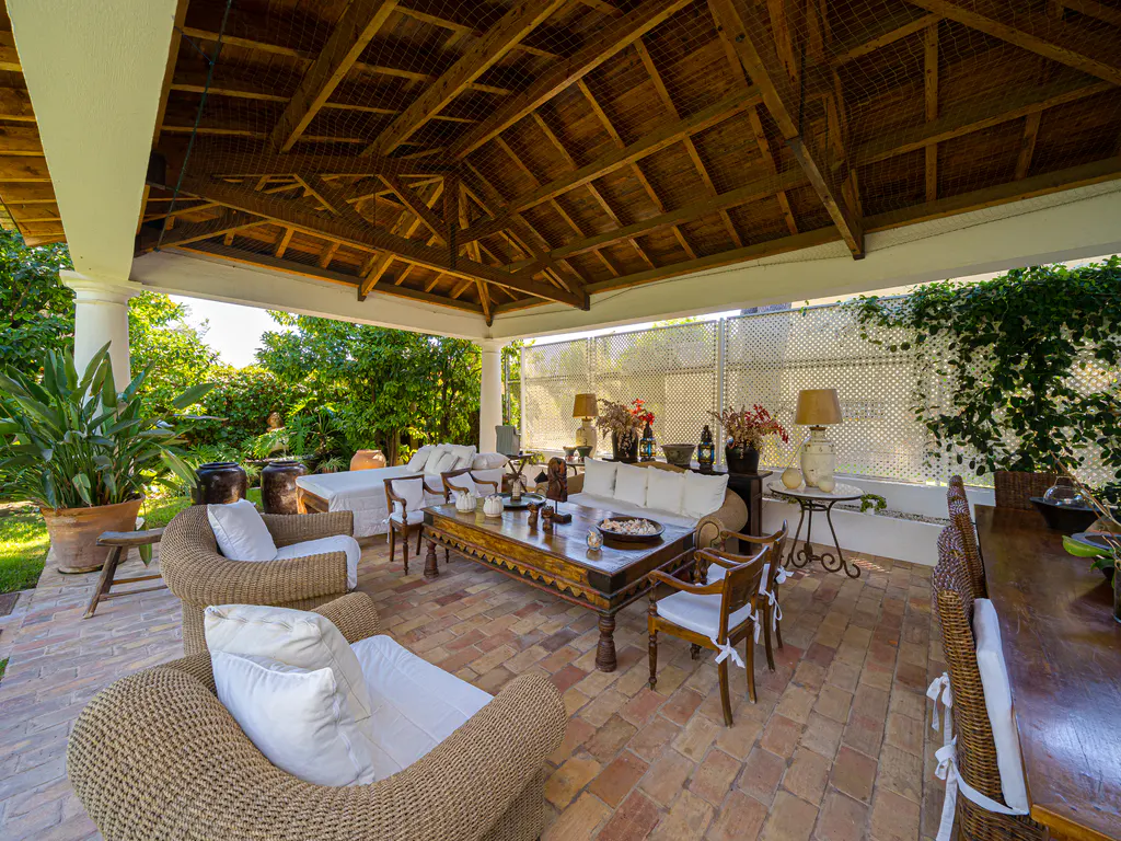 Covered outdoor patio with brick flooring, wicker furniture, a large wooden coffee table, and lush garden views.