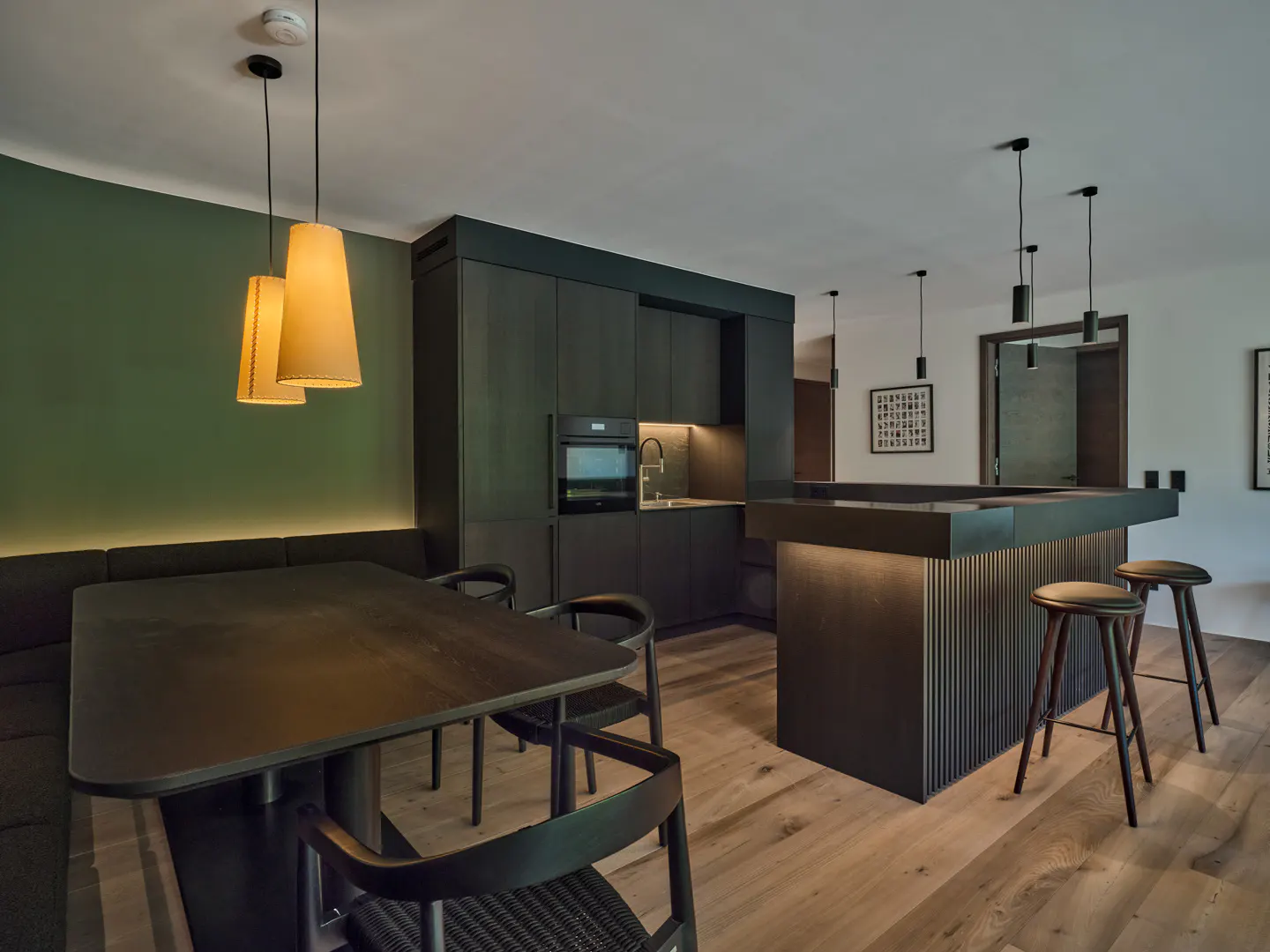 Modern kitchen with dark cabinetry, a dining table with bench seating, and a breakfast bar with stools on wood flooring.