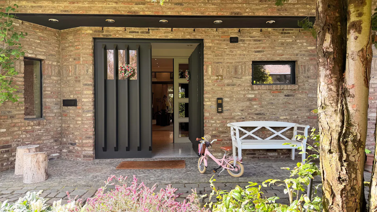 Brick house with an open black door, a pink bicycle, and a white bench on a stone porch.