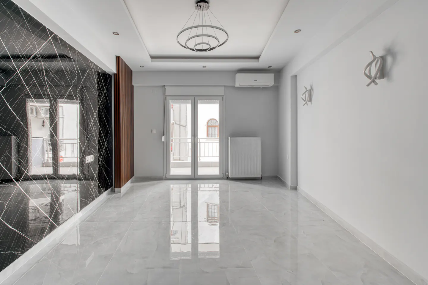Bright, empty room with marble floors, a black marble accent wall, and white walls. A modern chandelier hangs from the ceiling. Glass doors lead to a balcony.