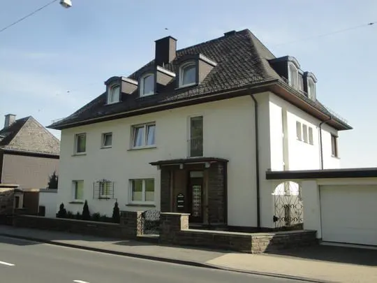 A two-story white house with a dark roof and dormer windows, seen from the street. A stone wall and gate surround the front yard.