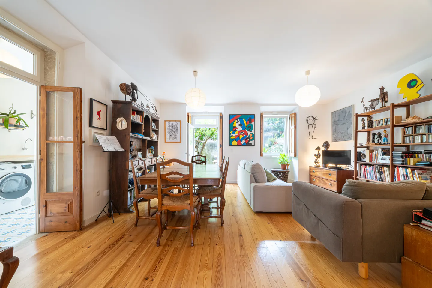 Bright living room with wood floors, dining table, sofas, bookshelves, and art. Natural light streams through windows.