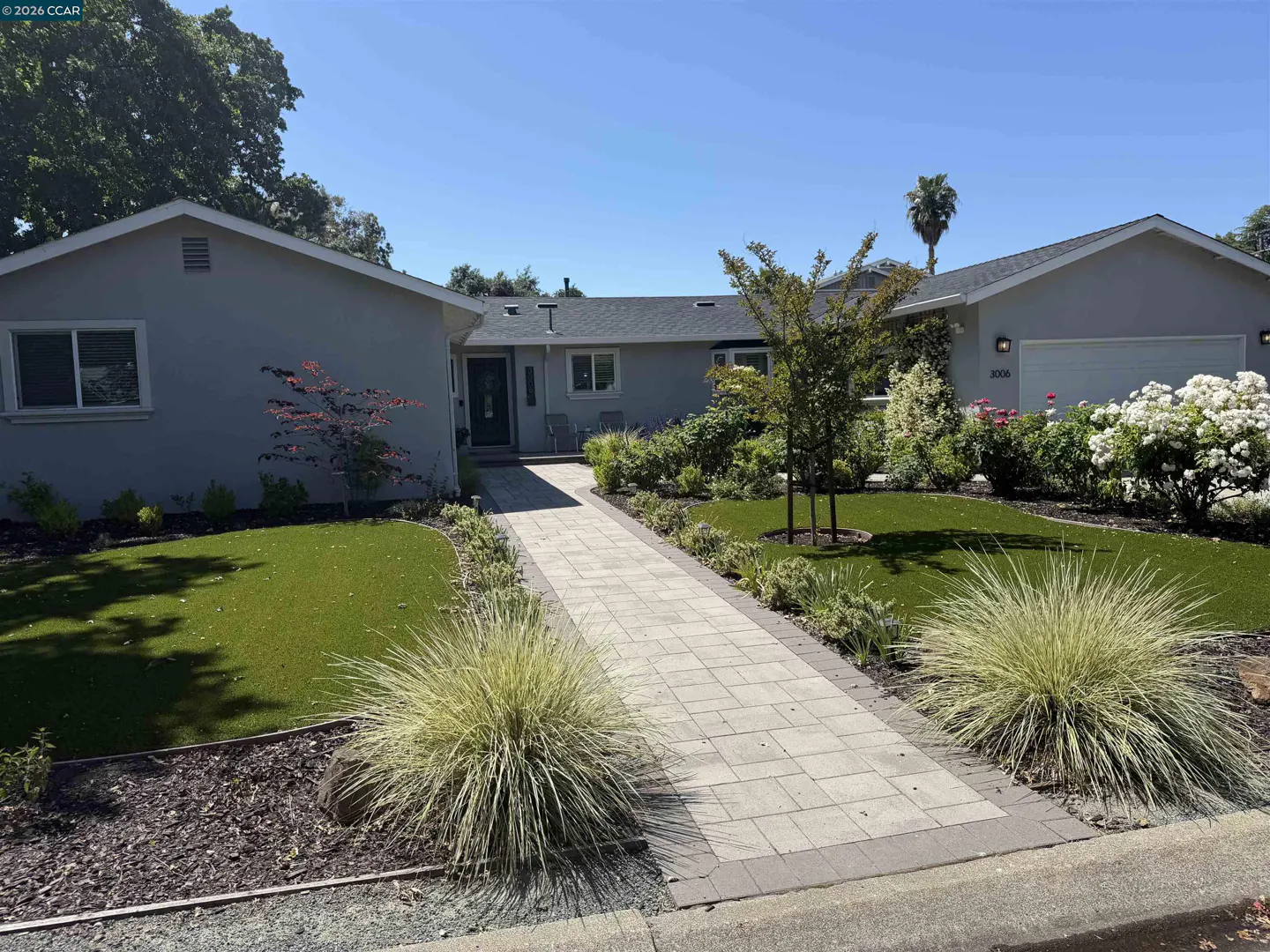 Exterior view of a one-story gray house with a stone walkway, green lawn, and lush landscaping on a sunny day.