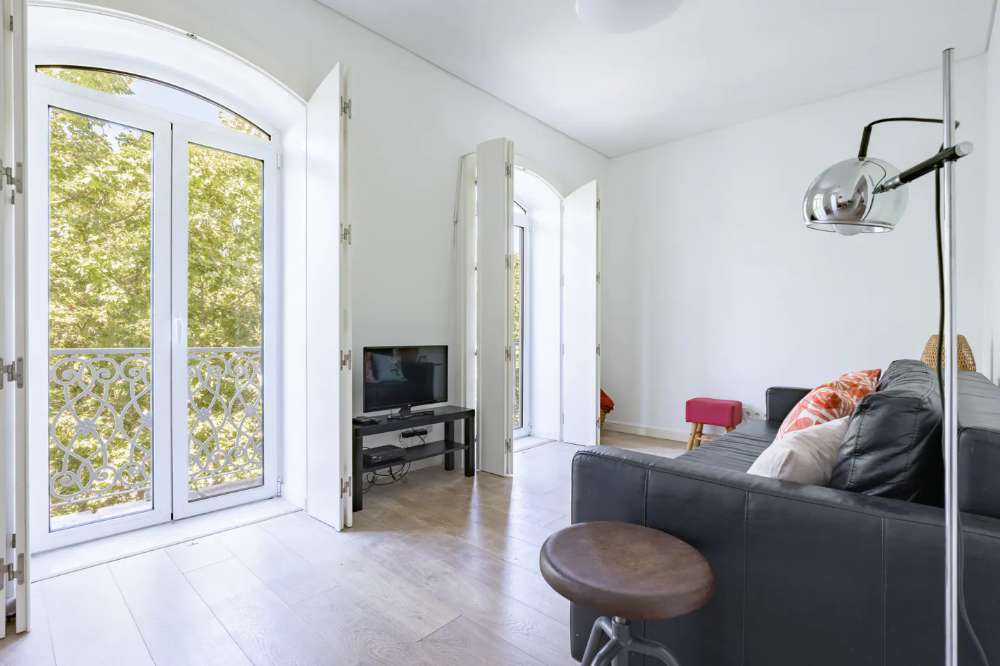 Bright living room with white walls, light wood floors, and a black leather sofa. White French doors open to a balcony with green trees.