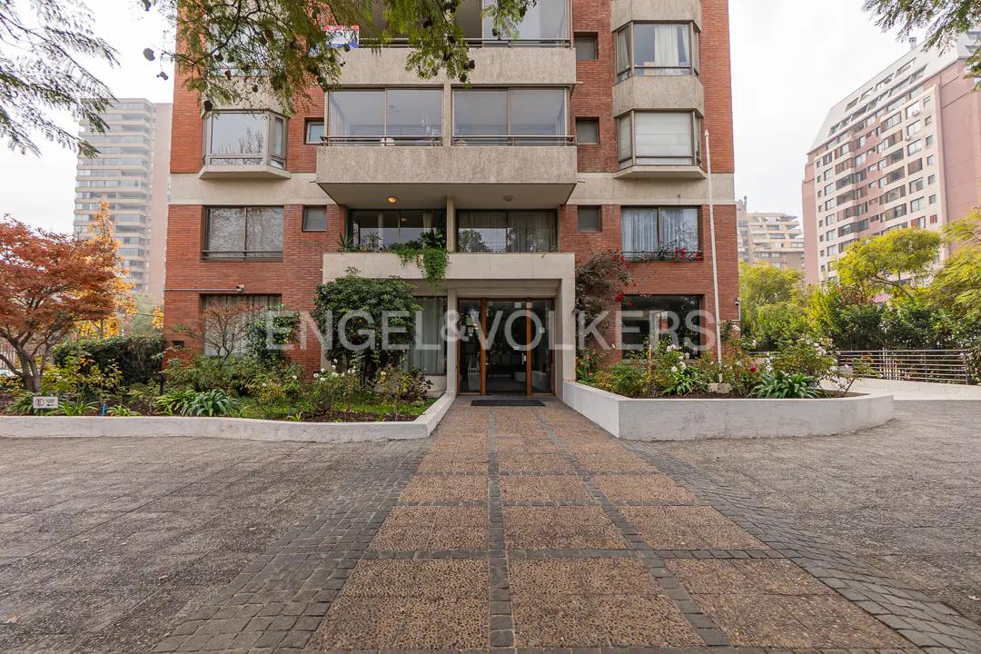 Entrance to a multi-story red brick apartment building with a paved walkway, surrounding greenery, and city buildings.