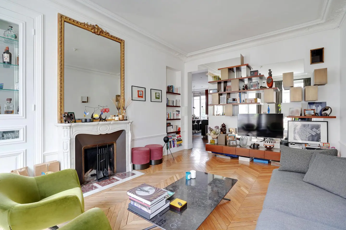 Bright living room with herringbone wood floors, fireplace with gold-framed mirror, green chairs, and a gray sofa.