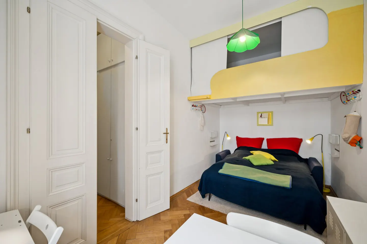A bedroom with a blue sofa bed, red pillows, and a yellow loft above. A green pendant lamp hangs from the ceiling. White closet doors are on the left.