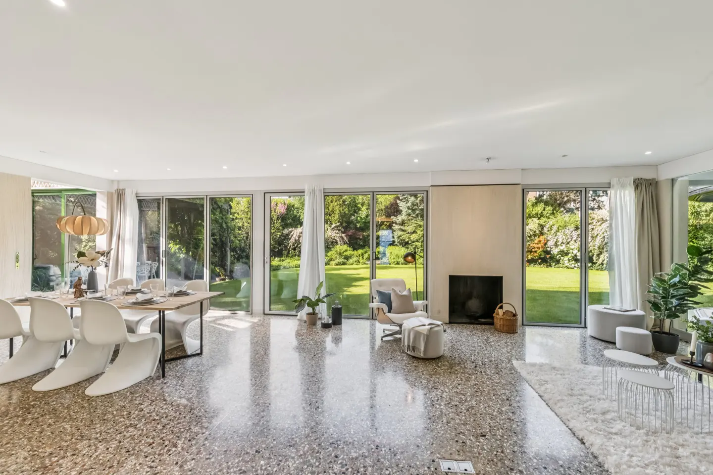 Bright, open-plan living and dining area with terrazzo floor. Dining table with white chairs, lounge with fireplace, and large windows overlooking a green garden.