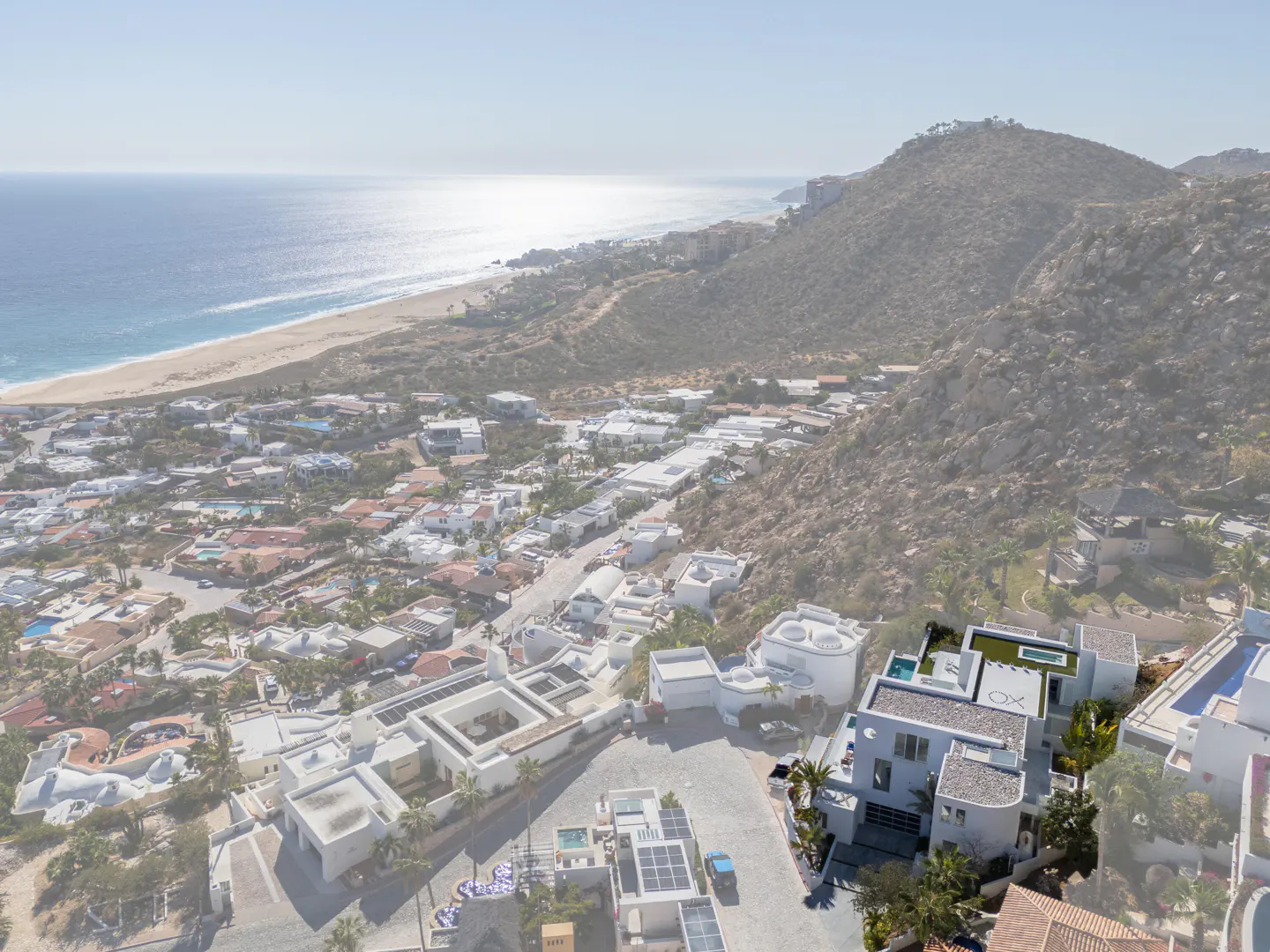 Aerial view of a modern white house with a rooftop pool, nestled on a hillside overlooking the ocean and a town.