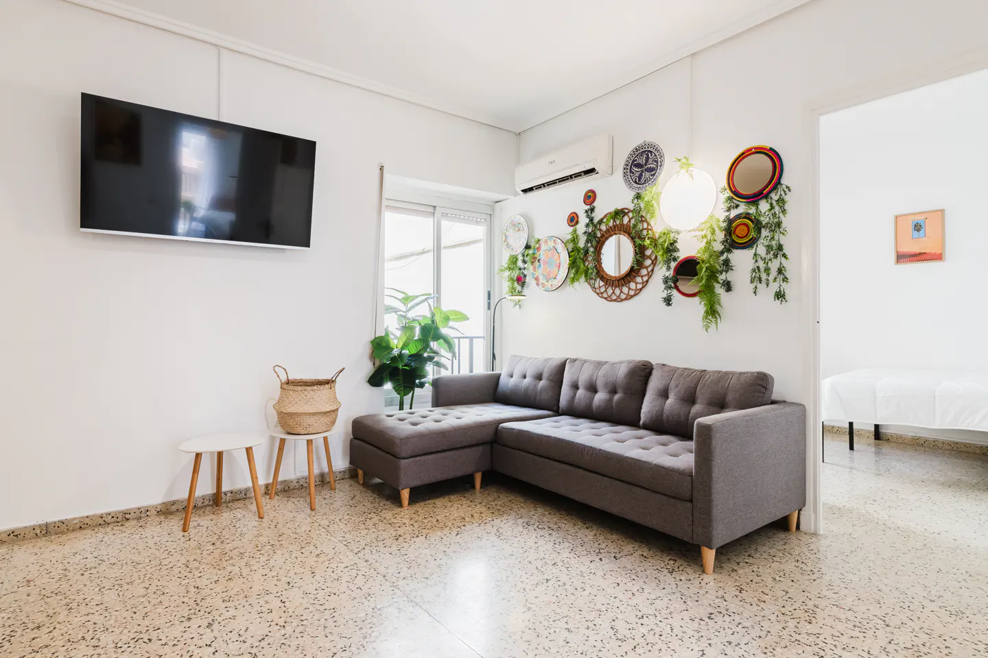 Living room with gray sectional sofa, TV, and decorative wall plates with greenery. White walls and terrazzo floor.