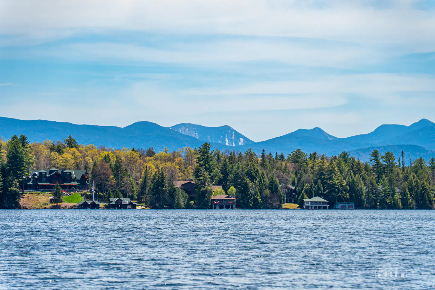 Lake view with houses nestled in trees along the shore, mountains in the background under a blue sky.