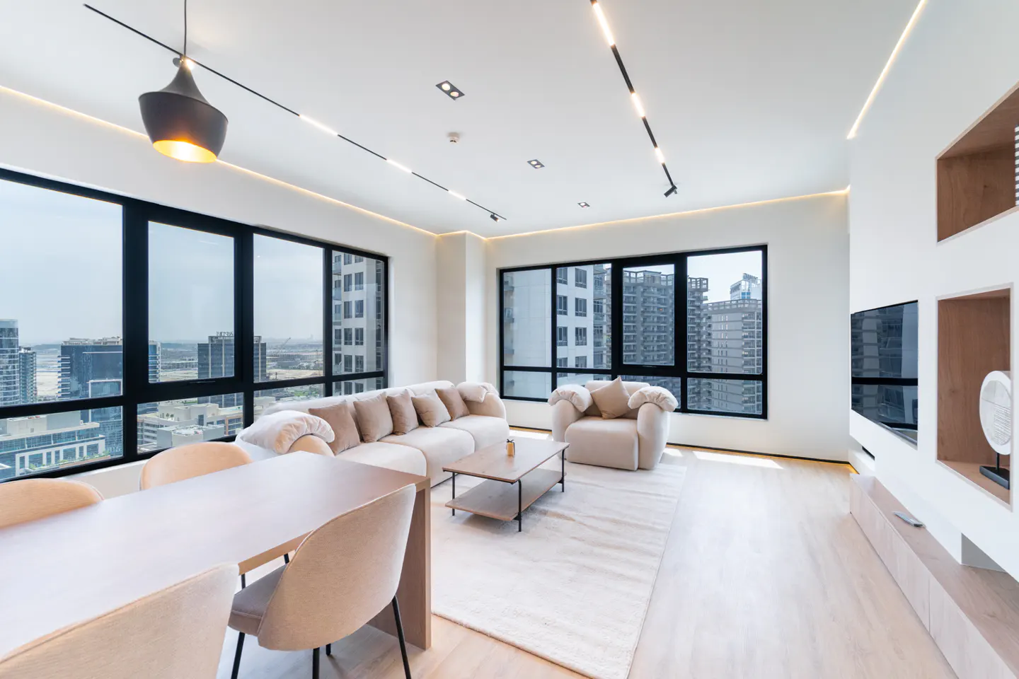 Bright, modern living room with beige sofa, chair, dining table, and city views through large windows.