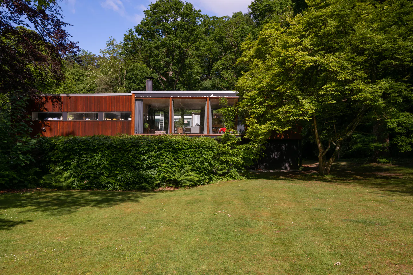 Exterior view of a modern, wood-clad home with large glass windows, surrounded by lush green trees and a lawn.