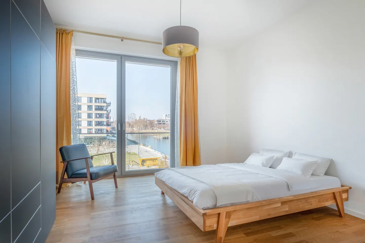 Bright bedroom with a wooden bed, white linens, and a blue chair by a large window with gold curtains overlooking a waterfront view.