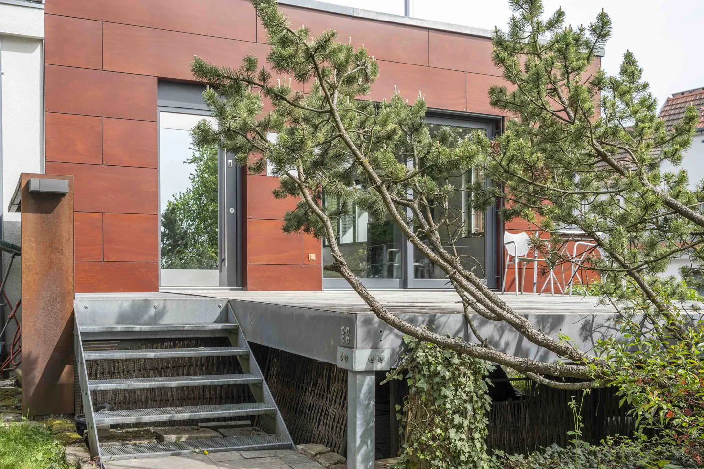 Exterior view of a modern home with a wood-paneled facade, a metal staircase leading to a deck, and a pine tree in the foreground.